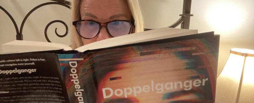 Woman with glasses holding the book Doppelganger by Naomi Klein open reading it